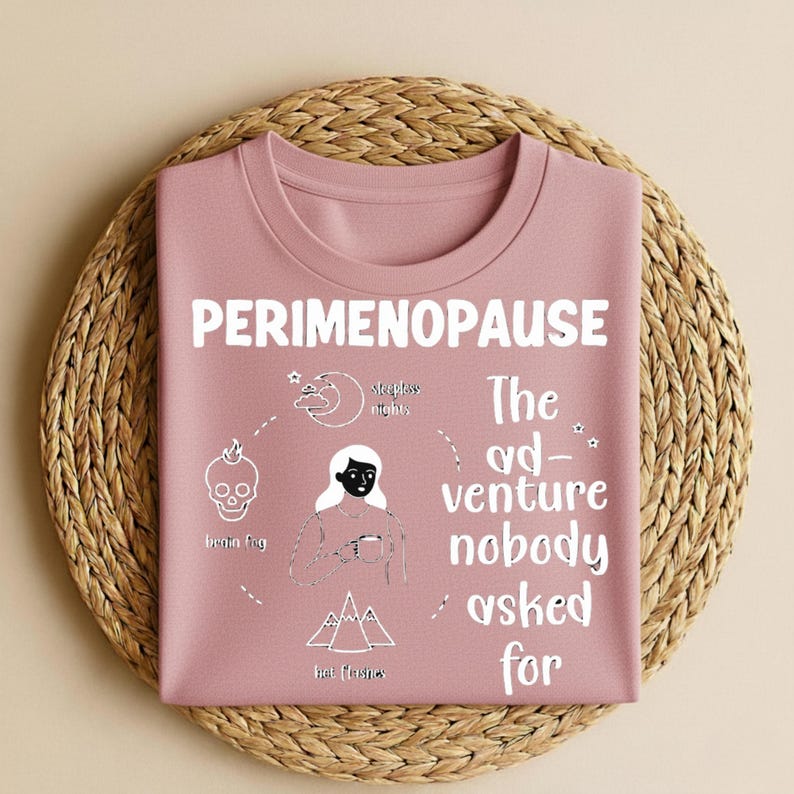 Puede incluir: Una sudadera de color rosa polvoriento con la palabra "PERIMENOPAUSE" en letras blancas. La camisa presenta un diagrama con ilustraciones de una calavera, una luna, monta&ntilde;as y una persona. El texto "The adventure nobody asked for" tambi&eacute;n est&aacute; en la camisa.