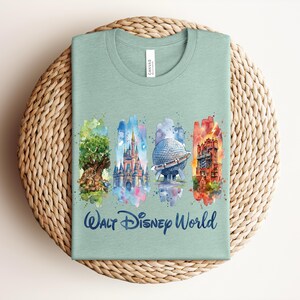 May include: A light green t-shirt featuring a watercolor design of Walt Disney World landmarks. The design includes the Tree of Life, Cinderella Castle, Spaceship Earth, and the Tower of Terror. The words "Walt Disney World" are printed in blue.