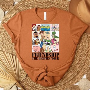 May include: Rust-colored t-shirt featuring a colorful graphic of cartoon characters. The graphic is divided into squares and includes the text "FRIENDSHIP THE BESTIES TOUR". The shirt has a crew neck and short sleeves.