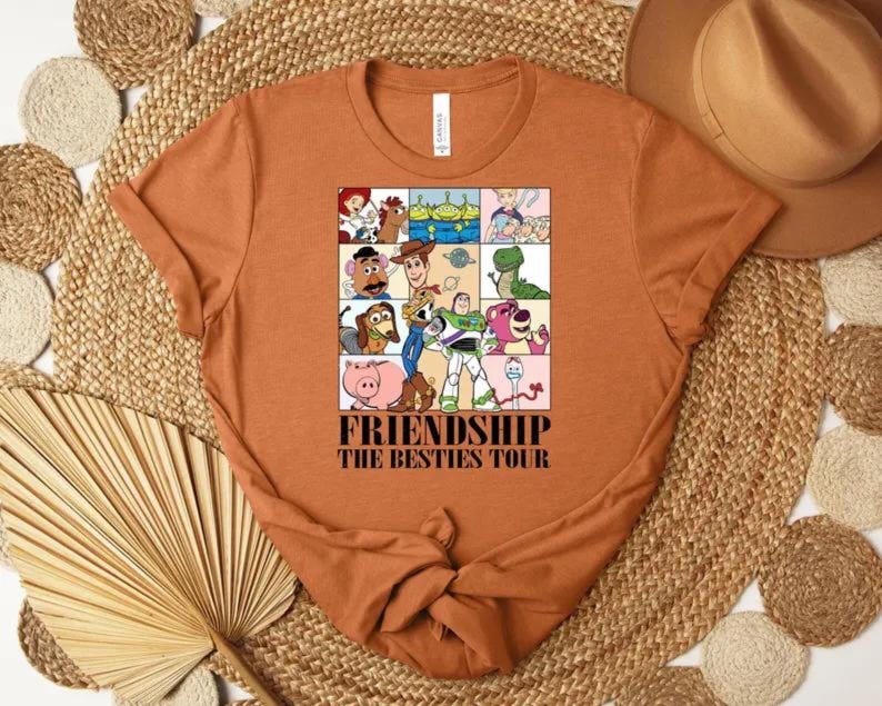 May include: Rust-colored t-shirt featuring a colorful graphic of cartoon characters. The graphic is divided into squares and includes the text "FRIENDSHIP THE BESTIES TOUR". The shirt has a crew neck and short sleeves.