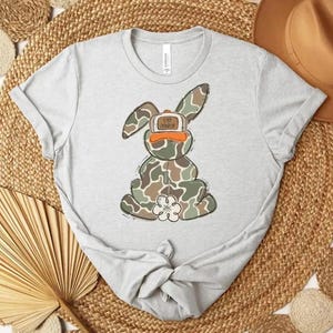 May include: A light gray t-shirt featuring a cartoon bunny in camouflage print. The bunny wears a hat with the text "UP NORTH". The shirt is styled on a woven mat with a hat and decorative elements.