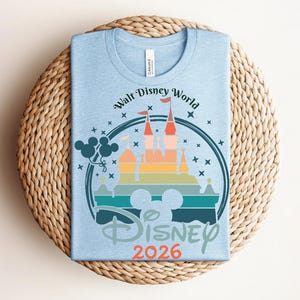 Op de afbeelding: Lichtblauw T-shirt met een afbeelding van een kasteel, een Mickey Mouse silhouet en de tekst "Walt Disney World Disney 2026". Het ontwerp toont een kleurrijk kasteel en sterren in een cirkelvormig kader.