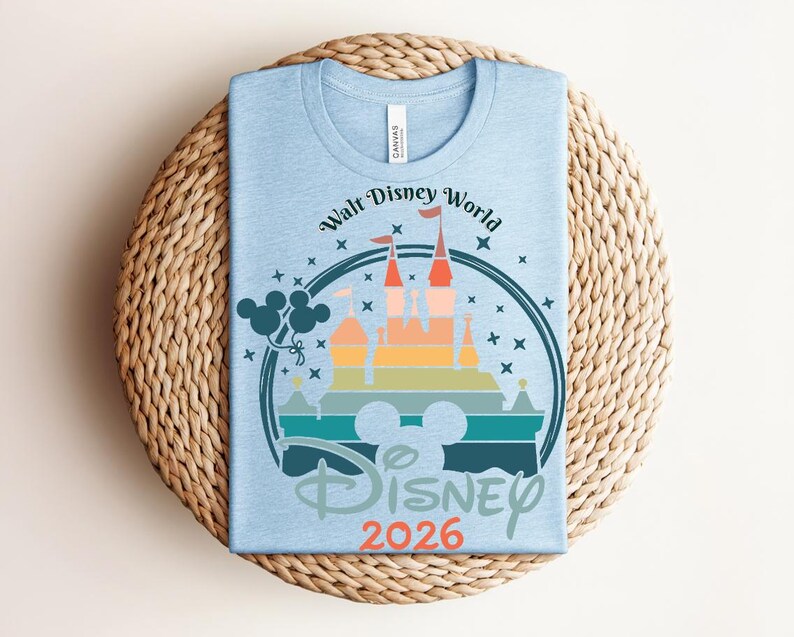 Op de afbeelding: Lichtblauw T-shirt met een afbeelding van een kasteel, een Mickey Mouse silhouet en de tekst "Walt Disney World Disney 2026". Het ontwerp toont een kleurrijk kasteel en sterren in een cirkelvormig kader.
