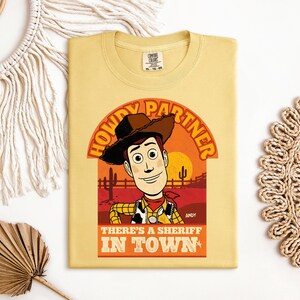 K&ouml;nnte beinhalten: Ein hellgelbes T-Shirt mit einer Cartoon-Illustration von Woody aus Toy Story. Das Design enth&auml;lt den Text "HOWDY PARTNER" und "THERE'S A SHERIFF IN TOWN". Die Grafik ist vor einem Sonnenuntergang mit Kakteen platziert.