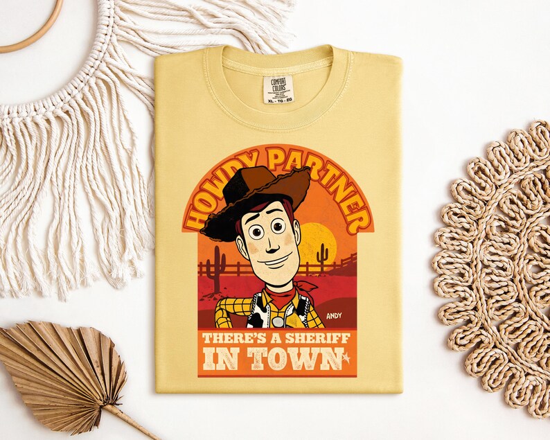 K&ouml;nnte beinhalten: Ein hellgelbes T-Shirt mit einer Cartoon-Illustration von Woody aus Toy Story. Das Design enth&auml;lt den Text "HOWDY PARTNER" und "THERE'S A SHERIFF IN TOWN". Die Grafik ist vor einem Sonnenuntergang mit Kakteen platziert.