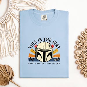 May include: A pale blue t-shirt with a graphic of a Mandalorian helmet and the phrase "THIS IS THE WAY". The design incorporates orange, yellow, and red elements. The words "BOUNTY HUNTER" and "CLAN OF TWO" are also printed on the shirt.