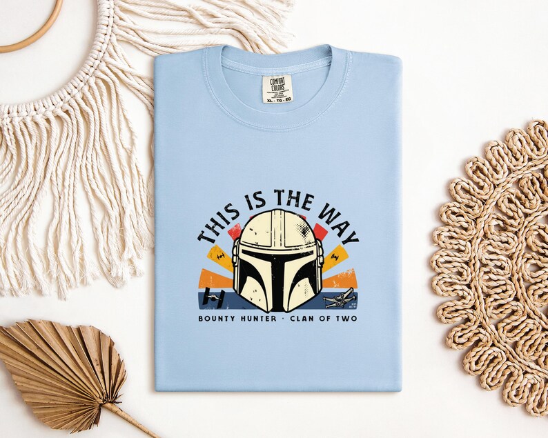 May include: A pale blue t-shirt with a graphic of a Mandalorian helmet and the phrase "THIS IS THE WAY". The design incorporates orange, yellow, and red elements. The words "BOUNTY HUNTER" and "CLAN OF TWO" are also printed on the shirt.