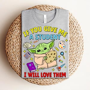 May include: A gray t-shirt with the text "IF YOU GIVE ME A STUDENT I WILL LOVE THEM." The shirt features a cartoon character holding a cookie, surrounded by school-related graphics like pencils, a calculator, and scissors.