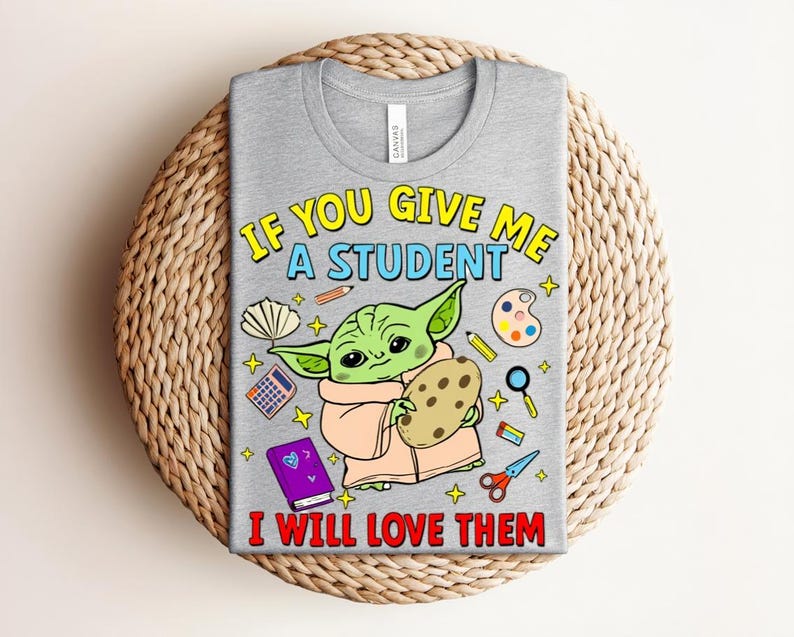 May include: A gray t-shirt with the text "IF YOU GIVE ME A STUDENT I WILL LOVE THEM." The shirt features a cartoon character holding a cookie, surrounded by school-related graphics like pencils, a calculator, and scissors.