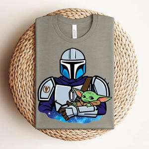 May include: A gray t-shirt featuring a cartoon illustration of a Mandalorian character holding a green alien. The Mandalorian is wearing a helmet and armor with blue accents. The alien is wrapped in a brown blanket, set against a starry background.
