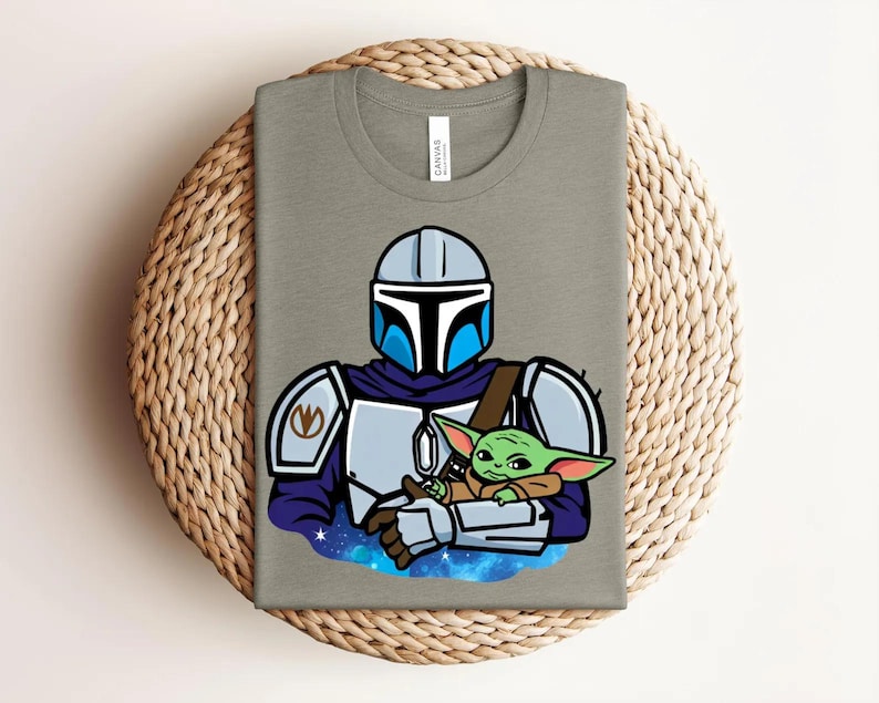 May include: A gray t-shirt featuring a cartoon illustration of a Mandalorian character holding a green alien. The Mandalorian is wearing a helmet and armor with blue accents. The alien is wrapped in a brown blanket, set against a starry background.