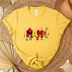 May include: Mustard yellow t-shirt with the word "MAMA" in colorful, comic-style lettering. The letters are decorated with superhero-themed graphics and accents, including hearts and stars. The shirt is folded and tied at the bottom.