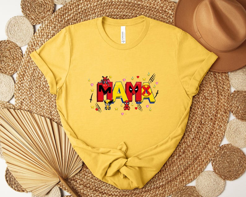 May include: Mustard yellow t-shirt with the word "MAMA" in colorful, comic-style lettering. The letters are decorated with superhero-themed graphics and accents, including hearts and stars. The shirt is folded and tied at the bottom.