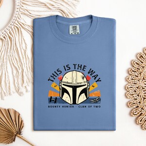 May include: A pale blue t-shirt with a graphic of a Mandalorian helmet and the phrase "THIS IS THE WAY". The design incorporates orange, yellow, and black details, with the words "BOUNTY HUNTER" and "CLAN OF TWO" beneath the helmet.