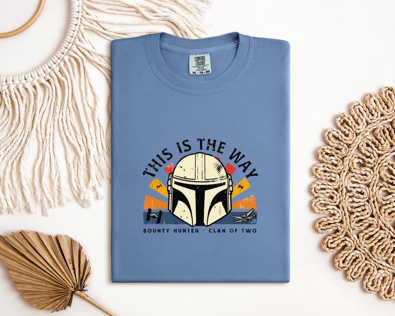 May include: A pale blue t-shirt with a graphic of a Mandalorian helmet and the phrase "THIS IS THE WAY". The design incorporates orange, yellow, and black details, with the words "BOUNTY HUNTER" and "CLAN OF TWO" beneath the helmet.
