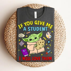 May include: Dark gray t-shirt featuring a cartoon character holding a cookie, surrounded by school supplies. The text reads, "IF YOU GIVE ME A STUDENT I WILL LOVE THEM" in yellow and red lettering. The shirt is displayed on a woven surface.