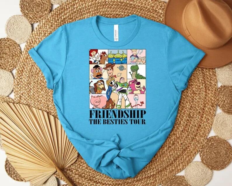 May include: A turquoise t-shirt featuring a colorful graphic of cartoon characters in a grid pattern. The graphic includes the text "FRIENDSHIP THE BESTIES TOUR" in black font. The shirt is made of a soft material.