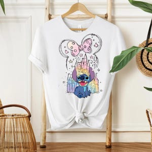 Pu&ograve; includere: T-shirt bianco con una grafica colorata del personaggio Stitch di Lilo & Stitch. Il design include un castello, stelle e un fiocco. La maglietta &egrave; annodata in basso.