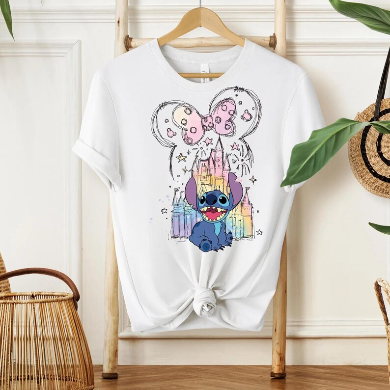 Pu&ograve; includere: T-shirt bianco con una grafica colorata del personaggio Stitch di Lilo & Stitch. Il design include un castello, stelle e un fiocco. La maglietta &egrave; annodata in basso.
