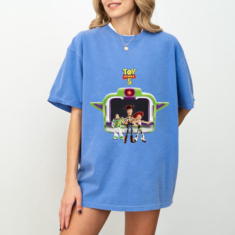 K&ouml;nnte beinhalten: Hellblaues T-Shirt mit einem Motiv aus dem Film Toy Story 5. Die Grafik zeigt Buzz Lightyear, Woody und Jessie in einem gr&uuml;n-violetten Rahmen. Das Toy Story 5-Logo befindet sich &uuml;ber dem Rahmen.