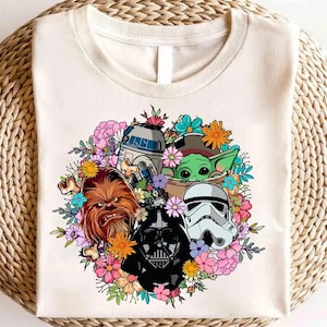 May include: Cream-colored t-shirt featuring a colorful floral wreath design with Star Wars characters. The graphic includes R2-D2, Chewbacca, Darth Vader, a Stormtrooper, and Grogu. The flowers are in shades of pink, blue, orange, and yellow.