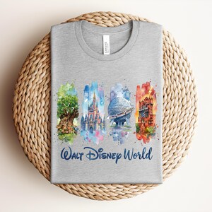 May include: A gray t-shirt featuring watercolor illustrations of iconic Walt Disney World landmarks: the Tree of Life, Cinderella Castle, Spaceship Earth, and the Tower of Terror. The words "Walt Disney World" are printed in blue script below the images.