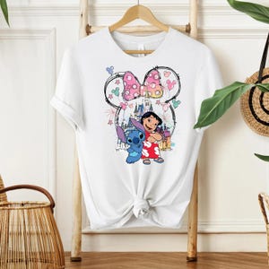 Pu&ograve; includere: T-shirt bianco con una grafica colorata di Lilo e Stitch, un fiocco rosa a pois e un castello. Il design include fuochi d'artificio e stelle, creando un'estetica giocosa. La maglietta &egrave; annodata in vita.
