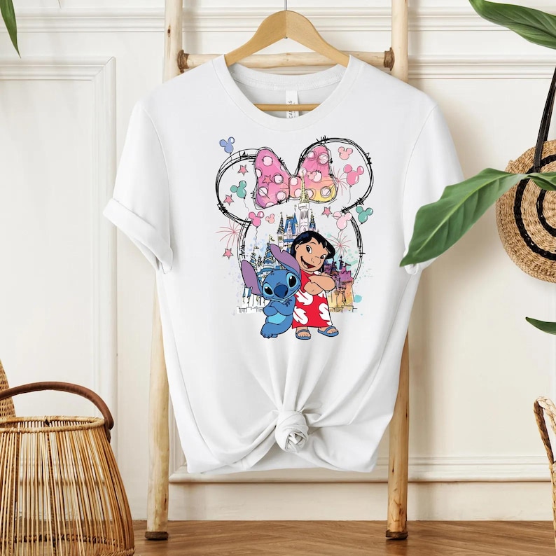 Pu&ograve; includere: T-shirt bianco con una grafica colorata di Lilo e Stitch, un fiocco rosa a pois e un castello. Il design include fuochi d'artificio e stelle, creando un'estetica giocosa. La maglietta &egrave; annodata in vita.