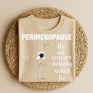 Puede incluir: Camiseta beige con la palabra "PERIMENOPAUSE" en letras blancas. La camiseta presenta un gr&aacute;fico de una mujer, una calavera, una luna y monta&ntilde;as. El texto "The ad-venture nobody asked for" tambi&eacute;n est&aacute; impreso.