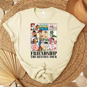 May include: Cream-colored t-shirt featuring a colorful graphic of cartoon characters from the movie Toy Story. The graphic is divided into squares and includes the text "FRIENDSHIP THE BESTIES TOUR".