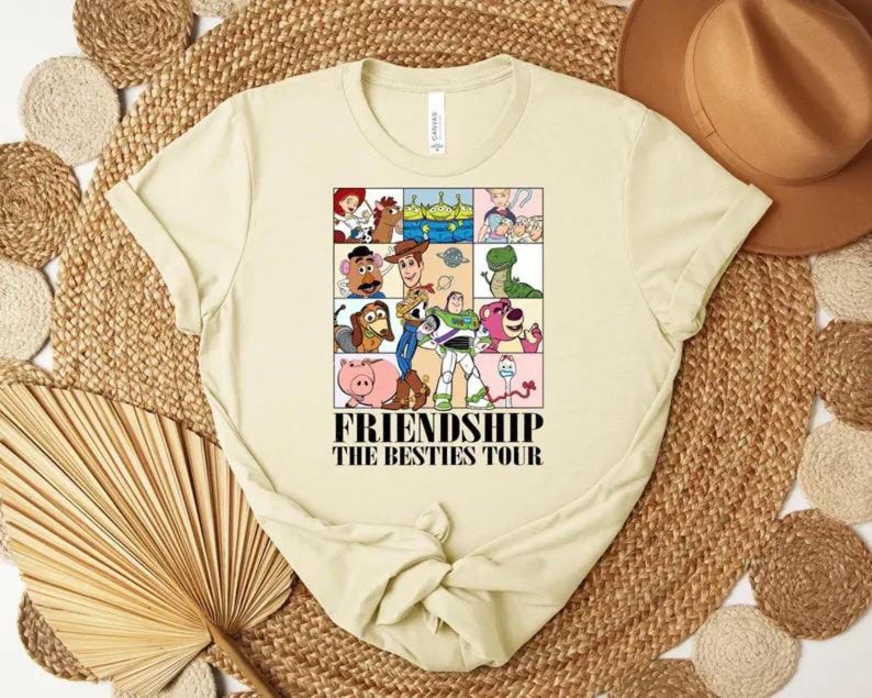 May include: Cream-colored t-shirt featuring a colorful graphic of cartoon characters from the movie Toy Story. The graphic is divided into squares and includes the text "FRIENDSHIP THE BESTIES TOUR".