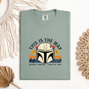 May include: A sage green t-shirt with a Mandalorian helmet graphic and the phrase "THIS IS THE WAY". The design incorporates orange, yellow, and blue elements, with "BOUNTY HUNTER" and "CLAN OF TWO" beneath. The t-shirt is folded neatly.