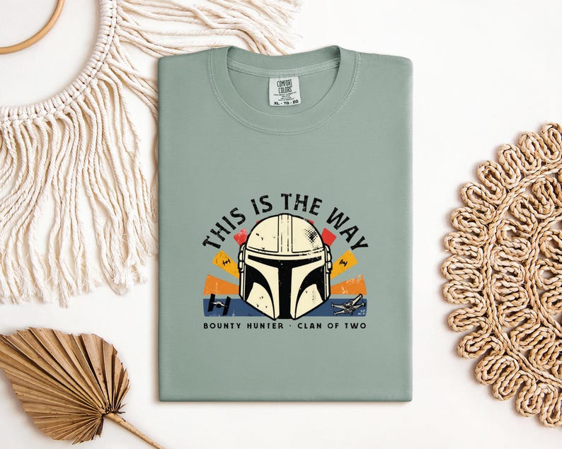 May include: A sage green t-shirt with a Mandalorian helmet graphic and the phrase "THIS IS THE WAY". The design incorporates orange, yellow, and blue elements, with "BOUNTY HUNTER" and "CLAN OF TWO" beneath. The t-shirt is folded neatly.