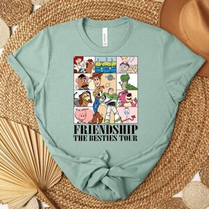 May include: A light green t-shirt featuring a colorful graphic of cartoon characters in a grid pattern. The graphic includes the text "FRIENDSHIP THE BESTIES TOUR" in a bold font. The shirt is folded, with the sleeves and bottom tied in a knot.
