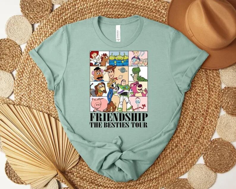 May include: A light green t-shirt featuring a colorful graphic of cartoon characters in a grid pattern. The graphic includes the text "FRIENDSHIP THE BESTIES TOUR" in a bold font. The shirt is folded, with the sleeves and bottom tied in a knot.