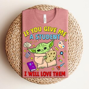 May include: A dusty rose t-shirt with the text "IF YOU GIVE ME A STUDENT I WILL LOVE THEM". The graphic features a green alien-like character holding a cookie, surrounded by school supplies. The shirt is displayed on a woven surface.