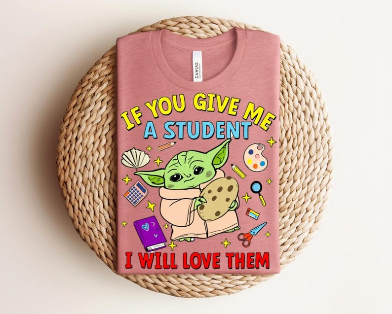 May include: A dusty rose t-shirt with the text "IF YOU GIVE ME A STUDENT I WILL LOVE THEM". The graphic features a green alien-like character holding a cookie, surrounded by school supplies. The shirt is displayed on a woven surface.