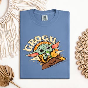 May include: A blue t-shirt featuring a Grogu graphic. The design includes the word "GROGU" in yellow, along with an illustration of Grogu holding a frog. The shirt is made of a soft material and is perfect for fans of the series.
