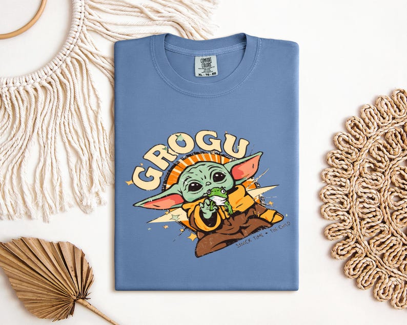 May include: A blue t-shirt featuring a Grogu graphic. The design includes the word "GROGU" in yellow, along with an illustration of Grogu holding a frog. The shirt is made of a soft material and is perfect for fans of the series.