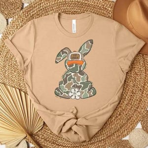 May include: A tan t-shirt featuring a cartoon bunny wearing a camouflage pattern and an orange cap. The bunny has a white flower on its chest. The shirt is knotted at the bottom and is displayed on a woven mat.