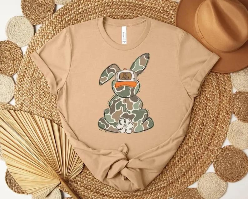 May include: A tan t-shirt featuring a cartoon bunny wearing a camouflage pattern and an orange cap. The bunny has a white flower on its chest. The shirt is knotted at the bottom and is displayed on a woven mat.