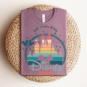 Op de afbeelding: Een mauvekleurig T-shirt met een kleurrijke afbeelding van een kasteel, Mickey Mouse-ballonnen en de tekst "Walt Disney World Disney 2026". Het ontwerp staat in een cirkelvormige lijst met sterren.