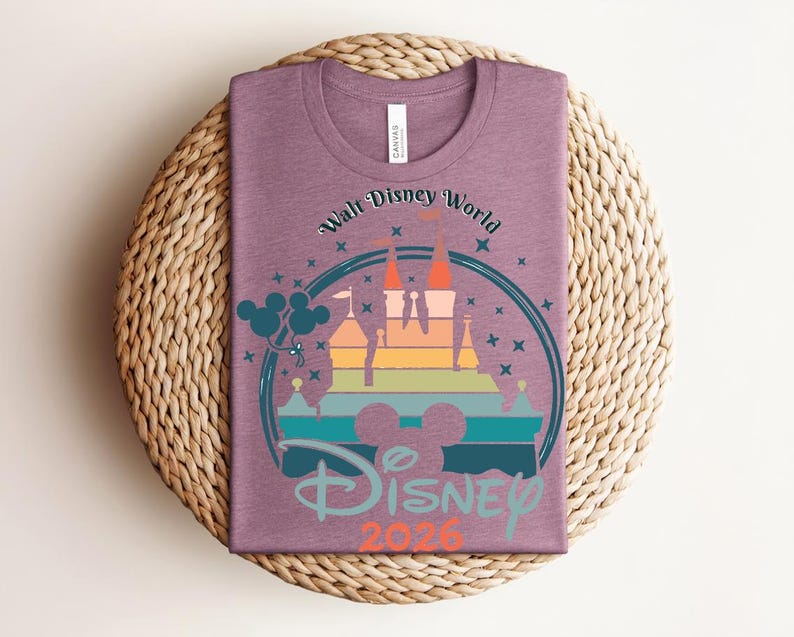 Op de afbeelding: Een mauvekleurig T-shirt met een kleurrijke afbeelding van een kasteel, Mickey Mouse-ballonnen en de tekst "Walt Disney World Disney 2026". Het ontwerp staat in een cirkelvormige lijst met sterren.