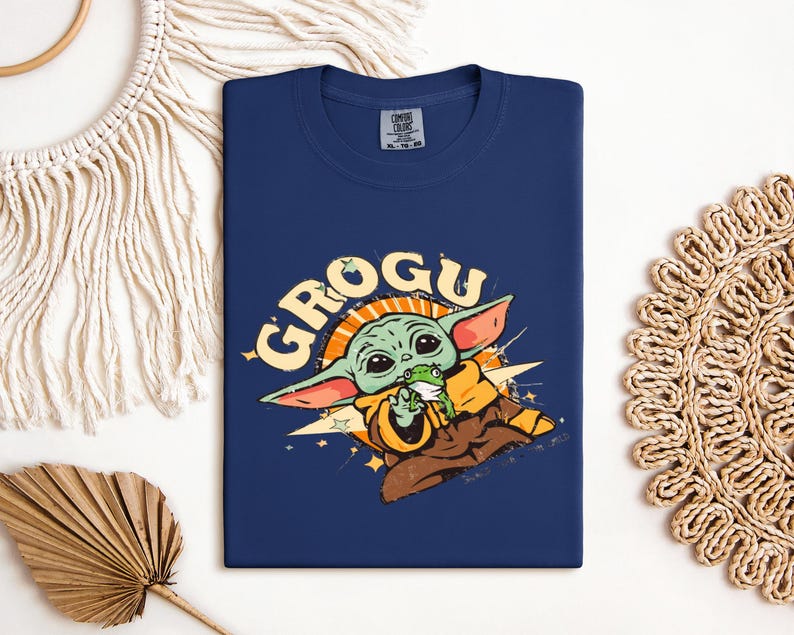 May include: Navy blue t-shirt featuring a Grogu graphic. The design includes Grogu holding a frog, with the word "GROGU" in a retro style. The shirt is a comfortable, casual wear item.