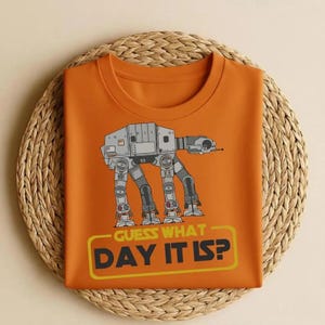 May include: Orange t-shirt featuring a graphic of an AT-AT walker from Star Wars. The design includes the text "GUESS WHAT DAY IT IS?" in a yellow rectangular box. The shirt is folded and placed on a woven, circular mat.