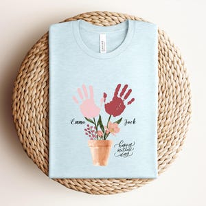 May include: Light blue t-shirt with a floral design featuring two handprints, one pink and one red, above a terracotta pot. The names "Emma" and "Jack" are printed below the handprints, with "happy mother's day" written in cursive.
