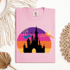 May include: A light pink t-shirt featuring a colorful graphic of a castle silhouette against a sunset backdrop. A fairy and a bow are also included in the design. The shirt has a crew neck and a tag on the collar.