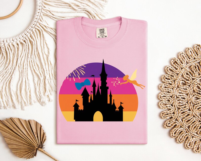 May include: A light pink t-shirt featuring a colorful graphic of a castle silhouette against a sunset backdrop. A fairy and a bow are also included in the design. The shirt has a crew neck and a tag on the collar.