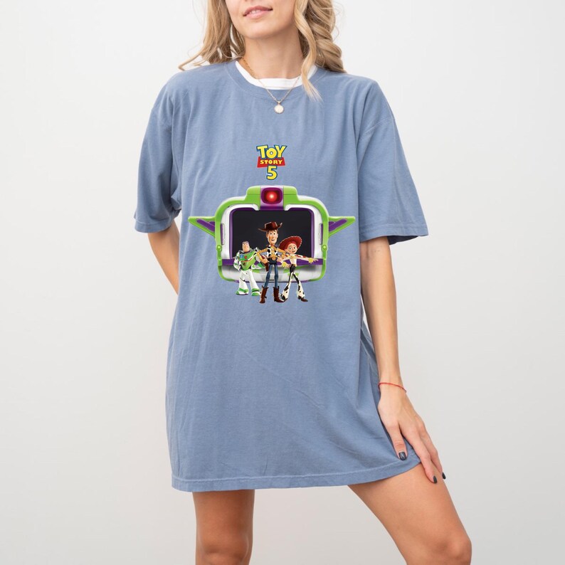 K&ouml;nnte beinhalten: Blaues T-Shirt-Kleid mit einem Aufdruck aus dem Film Toy Story 5. Der Aufdruck zeigt Woody, Buzz Lightyear und Jessie, mit dem Text "Toy Story 5" dar&uuml;ber. Das Kleid hat eine einfarbige blaue Farbe.