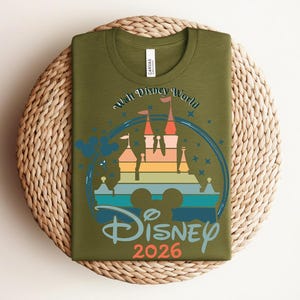Op de afbeelding: Olijfgroen T-shirt met een kleurrijke afbeelding van een kasteel, Mickey Mouse oren en de woorden "Walt Disney World" en "Disney 2026". Het ontwerp bevat blauwe, oranje, gele en groene elementen.